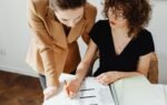 Two women discussing and analyzing business paperwork together in an office setting.