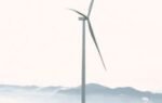 A wind turbine stands tall against mountains and farm fields, symbolizing sustainable energy.