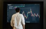 Back view of a trader analyzing cryptocurrency data on a monitor indoors.