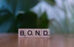 Scrabble tiles spelling 'Bond' on a wooden table with a blurred green background.