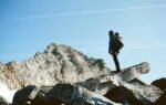 Full body of anonymous person on rocky rough formation among mountains under bright blue sky with airplane cloud